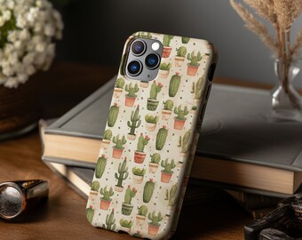 Puede incluir: Una funda blanca para iPhone con un patrón de cactus verdes en macetas de terracota. La funda tiene un recorte para el objetivo de la cámara y un recorte para los botones del teléfono.