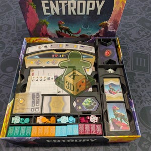 Può includere: La scatola del gioco da tavolo Entropy è aperta, rivelando un inserto in plastica nera che contiene vari componenti di gioco. Include mazzi di carte, piccoli gettoni colorati, tessere rettangolari e una plancia giocatore. Il coperchio riporta il titolo ENTROPY.