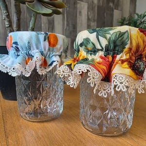 May include: Two clear glass jars with patterned fabric covers. One cover features orange fruit on a light blue background, the other has a floral design with yellow and red hues. Both covers have white lace trim.