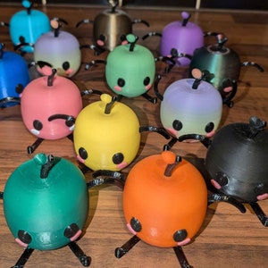 Puede incluir: Una colección de 12 pequeñas figuritas coloridas impresas en 3D. Cada figurita tiene forma de esfera con patas cortas negras y una pequeña antena negra en la parte superior. Las figuritas están dispuestas en círculo sobre una superficie de madera.