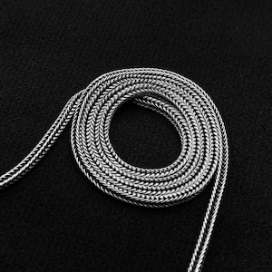 May include: A coiled silver-colored chain necklace against a black background. The chain is made of small, tightly woven links, creating a textured appearance. The necklace is in a circular pattern.