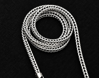 May include: A silver-toned wheat chain necklace featuring a tightly woven, braided metal link pattern. The chain is coiled into a circular shape, showcasing the intricate, repeating texture of the metallic links and a polished finish.