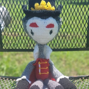 May include: A crocheted plush toy in the shape of a vampire with a black hat and a yellow crown. The toy is wearing a red and yellow waistcoat and has a white face with red eyes.