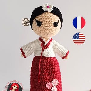 Peut inclure: Une poupée au crochet portant une robe traditionnelle coréenne hanbok. La poupée a les cheveux noirs, une fleur blanche dans les cheveux et une robe rouge et blanche. La poupée est debout sur un fond blanc.