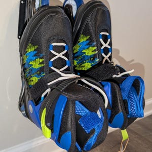 May include: Pair of black inline skates with blue and green accents. The skates have white laces, blue wheels, and a blue and black design. The skates are hanging on a wall.