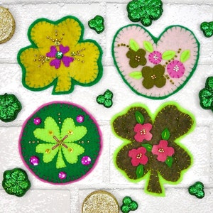 May include: Four felt shamrock ornaments with different designs. One is yellow with pink and green beads, one is green with pink beads, one is pink with green leaves and pink flowers, and one is brown with green leaves and pink flowers.