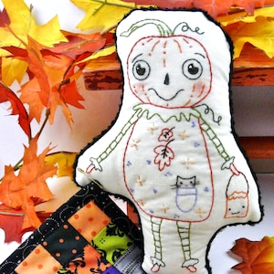 May include: A handmade Halloween-themed doll with a pumpkin face, wearing a white dress with embroidered details, holding a small orange bucket. The doll is sitting on a patchwork quilt with black, orange, green, and purple squares.