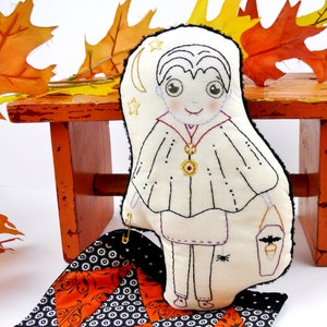 May include: A handmade Halloween-themed doll with a vampire design, featuring a cream-colored cape and a small bucket. The doll is placed on a patterned orange and black fabric, with autumn leaves in the background. The doll is approximately 8 inches tall.