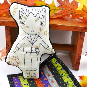 May include: A handmade plush toy of a Frankenstein monster holding a pumpkin bucket. The toy is dressed in a white shirt and pants and has stitched-on details. The toy is sitting on a patchwork quilt with a Halloween theme.