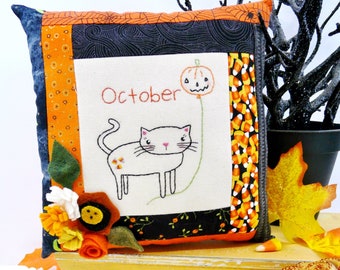 Op de afbeelding: Een handgemaakt kussen met een geborduurde kat en het woord "Oktober" op een witte achtergrond. Het kussen is versierd met oranje, zwart en wit stof en heeft een viltbloemversiering.