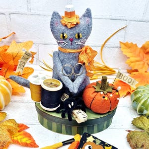 Op de afbeelding: Grijze vilten kattenpop met hoed en oranje sjaal, met een schaar. Vilten pompoen, spin en borduurgaren op een ronde doos. Versierd met herfstbladeren en een banner met "Boo!"