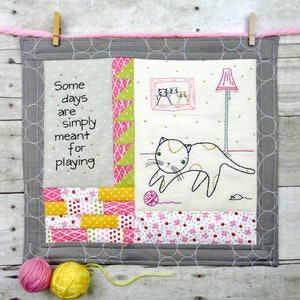 May include: A gray and white quilted wall hanging with a cat playing with a ball of yarn. The text "Some days are simply meant for playing" is stitched on the left side of the hanging.