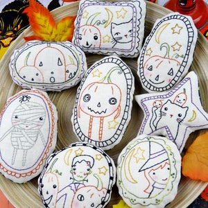 May include: Eight handmade Halloween-themed embroidered ornaments in a variety of shapes and designs, including pumpkins, cats, a witch, a mummy, and a Frankenstein monster. The ornaments are displayed on a wooden tray with autumn leaves.