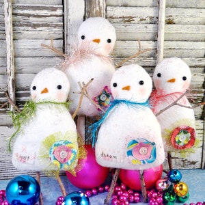 Puede incluir: Cuatro adornos de muñeco de nieve de fieltro blanco con detalles en rosa, azul y verde. Cada muñeco de nieve tiene una palabra diferente bordada en la parte delantera: "esperanza", "sueño" y dos sin texto. Los muñecos de nieve están rodeados de cuentas rosas y adornos de vidrio azul y verde.