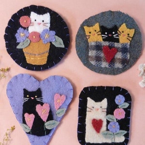 May include: Four felt cat ornaments with different designs. One features a white cat in a flower pot, another has three cats in a pocket, one has a black cat with hearts, and the last has a white cat with a heart.