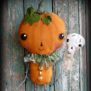 May include: A handmade pumpkin doll with a friendly face and green leaves on its head. The doll is wearing a green fabric collar and has twig legs. A white fabric ghost doll with a stitched-on face is standing next to the pumpkin doll. Both dolls are displayed against a weathered green wooden background.
