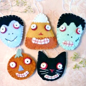 May include: Five felt Halloween decorations, each featuring a different character: a vampire, a candy corn, a Frankenstein, a pumpkin, and a black cat. Each decoration has a stitched-on face with googly eyes.