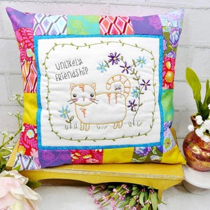 Puede incluir: Cojín decorativo cuadrado con un diseño de patchwork multicolor. El centro presenta un bordado de un gato y un ratón con las palabras "Unlikely Friendship". El cojín está colocado sobre una superficie amarilla.