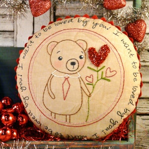 Puede incluir: Una almohada de tela beige en forma de corazón con texto bordado que dice "¡Quiero ser amado por ti! ¡Quiero ser amado por ti!"  La almohada presenta un oso de peluche marrón sosteniendo un corazón rojo de cuentas en un tallo verde con dos corazones más pequeños.