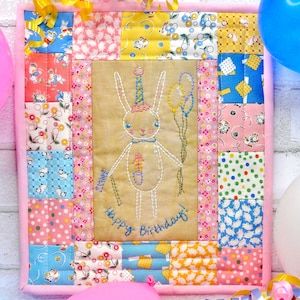 May include: A pink and yellow patchwork quilt with a bunny embroidered in the center. The bunny is wearing a party hat and holding balloons. The quilt says "Happy Birthday!" in a cursive font.