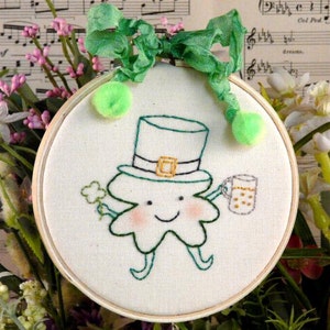 May include: Embroidered hoop art featuring a green shamrock with a smiling face, wearing a green hat and holding a pint of beer. The shamrock is surrounded by a white background and a wooden hoop.