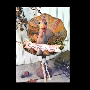Op de afbeelding: Een handgemaakt Thanksgiving-kalkoenbeeldje met bruine en oranje gepatroneerde staartveren. De kalkoen zit op een voetstuk met een banner waarop "Happy Thanksgiving!" staat.