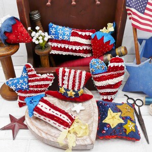 May include: A selection of red, white, and blue fabric ornaments with a patriotic theme. The collection includes star, flag, and heart shapes, a top hat, and a strawberry, all decorated with lace and embroidery.