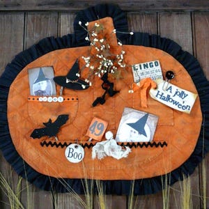 Op de afbeelding: Een gewatteerde oranje pompoenvormige wandhanger met een zwarte ruchesrand. Het bevat zakken met een vintage foto, een zwarte heksenhoed, een vleermuisfiguur, een bingokaart en labels met de tekst A jolly Halloween en het getal 49.