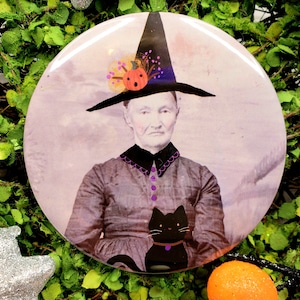 May include: A round Halloween-themed button featuring a vintage portrait of a woman wearing a witch hat and holding a black cat. The button has a light pink background and is surrounded by green foliage and silver stars.
