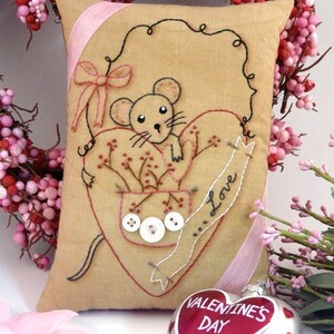 May include: A beige pillow with a pink ribbon and a heart-shaped design. The heart features a mouse with a red nose and the words "Love" and "Valentine's Day".
