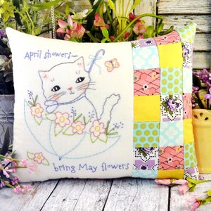 May include: White pillow with embroidered cat holding an umbrella and flowers. The text "April showers... bring May flowers" is embroidered on the pillow. The back of the pillow is made of patchwork fabric in yellow, pink, green, and purple.