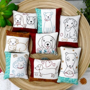 May include: Nine embroidered pillow covers with different dog designs. The pillows are all white with colorful accents and feature various dog breeds, including a poodle, dachshund, and labrador.