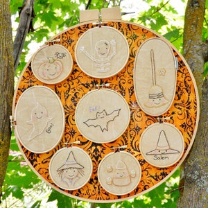 May include: A collection of nine embroidered Halloween-themed hoop art pieces. The hoops are arranged in a circle on a brown and orange background. The designs include a ghost, a bat, a pumpkin, a skeleton, a witch, a broom, a candy corn, and a spider.