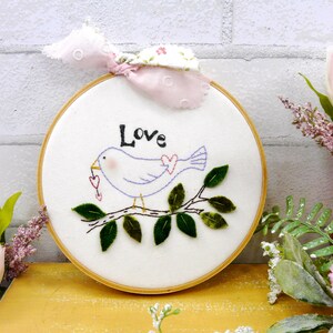 Könnte beinhalten: Gestickte Stickrahmenkunst mit einem hellvioletten Vogel, der ein Herz hält und auf einem Zweig mit grünen Blättern sitzt. Das Wort "Love" ist über dem Vogel gestickt. Der Rahmen ist in einem Holzring gefasst, mit einer rosa-weißen Stoffschleife oben.