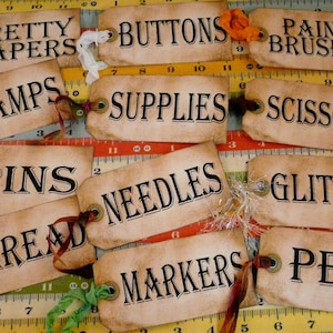 May include: Twelve vintage-style paper tags with brown edges and black text. The tags are labeled with craft supply terms like "Pretty Papers", "Buttons", "Paint Brushes", "Stamps", "Supplies", "Scissors", "Pins", "Needles", "Glitter", "Thread", "Markers", and "Pens".