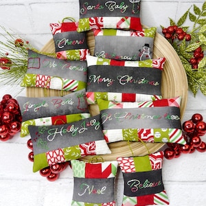 Puede incluir: Un conjunto de diez adornos navideños hechos de tela con diferentes lemas navideños bordados. Los adornos son de varios tonos de verde, rojo y gris con costuras blancas. Los adornos están dispuestos en un plato de madera con bayas rojas y verdes en el fondo.