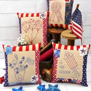 May include: A collection of patriotic throw pillows with embroidered designs. Pillows feature red, white, and blue color schemes with stars, stripes, balloons, and fireworks. A small American flag is also present.