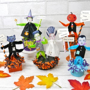 Puede incluir: Un conjunto de decoraciones de Halloween con una bruja, Frankenstein, un fantasma, una calabaza y un vampiro. Cada figura está hecha de papel y limpiapipas y sostiene un cartel con un mensaje de Halloween. Las figuras están sentadas sobre una variedad de bases, incluyendo una base negra y naranja, una base morada y verde y una base azul y morada.