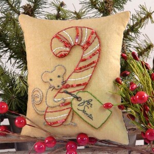 Pode incluir: Uma almofada bege com um desenho bordado de um rato segurando um bastão de caramelo com uma etiqueta que diz "Para o Papai Noel".