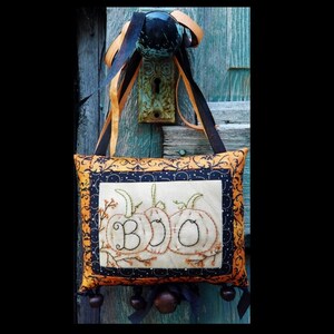 Puede incluir: Una almohada de Halloween hecha a mano con un diseño de tela negro y naranja. La almohada presenta un diseño bordado de tres calabazas con la palabra "BOO" escrita en las calabazas.