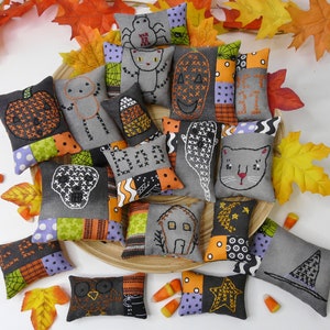 May include: A collection of 15 handmade Halloween-themed cushion ornaments. The cushions are made of patchwork fabric in shades of grey, black, orange, and white. Each cushion features a different embroidered design, including a pumpkin, a ghost, a cat, a skull, a spider, a witch's hat, a house, a star, and a candy corn.