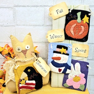 May include: A handmade seasonal banner set featuring a plush cat figure and four felt banners. The banners display a pumpkin, snowman, bunny, and crow, each representing a season. The tags read "Fall", "Winter", "Spring", and "Summer".