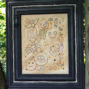 May include: A framed embroidery featuring a variety of Halloween-themed characters, including a pumpkin, a ghost, a spider, and a bat. The embroidery is stitched in black and white thread on a beige fabric.