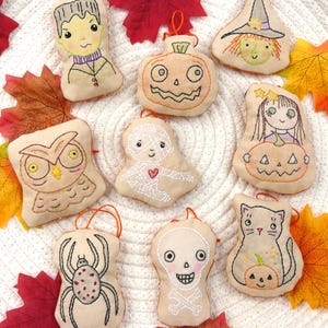 May include: A collection of eight beige fabric ornaments featuring embroidered Halloween designs including a monster, pumpkin, witch, owl, mummy, girl with pumpkin, spider, and skull with crossbones, each with an orange hanging loop.