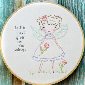 Op de afbeelding: Borduurwerk in een hoepel met een fee met bruin haar, een paarse jurk en vleugels. De fee houdt een roze bloem vast. De tekst "Little joys give us our wings" is geborduurd op de hoepel.