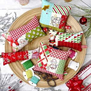 May include: A collection of handmade Christmas-themed pincushions in various shapes and sizes, arranged on a wooden tray. The pincushions feature festive patterns, including Santa Claus, snowmen, and geometric designs. Spools of thread and scissors are also present.