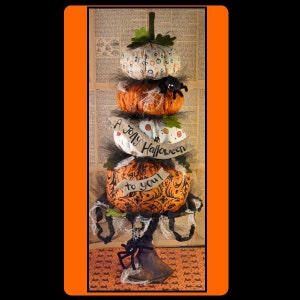May include: A stack of four fabric pumpkins decorated with black and white patterns, orange and black swirls, and a banner that reads "A Jolly Halloween to you!" The pumpkins are on a metal stand with a spider web and a black spider.