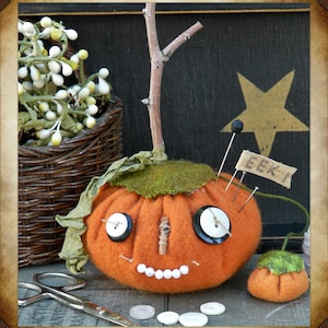 May include: A handcrafted orange felt pumpkin with button eyes, a twig nose, and a stitched smile. A small banner reads "EEK!" and is pinned to the pumpkin. A smaller pumpkin and scissors are also visible, suggesting a fall or Halloween theme.