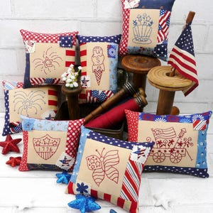 May include: A collection of patriotic throw pillows with red, white, and blue patterns and embroidered designs. Pillows feature fireworks, ice cream cones, butterflies, and the American flag. Decorative stars and a small American flag are also present.