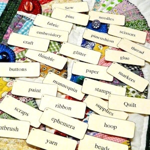 Puede incluir: Una vista cenital muestra una colección de tarjetas de palabras relacionadas con la artesanía, esparcidas sobre un edredón colorido. Las tarjetas, con una estética vintage, presentan términos como "tela", "tijeras", "hilo" y "botones". El diseño de patchwork del edredón proporciona un fondo vibrante.